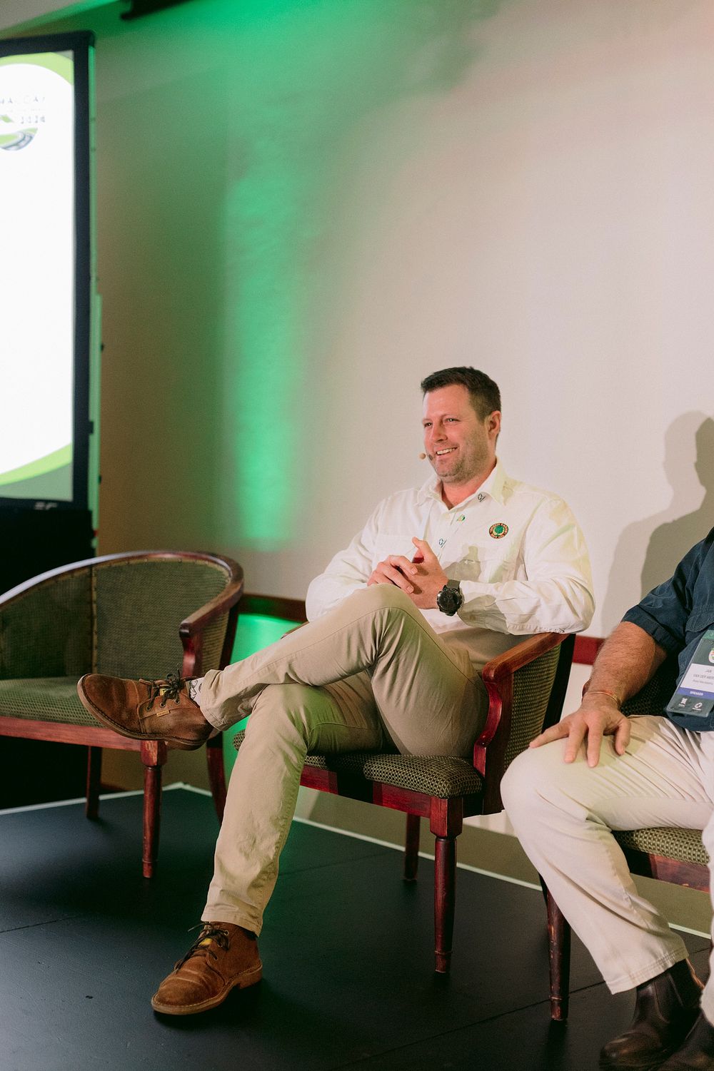 Man in white shirt sits smiling on stage panel, green lit backdrop, crossed legs, casual business attire.