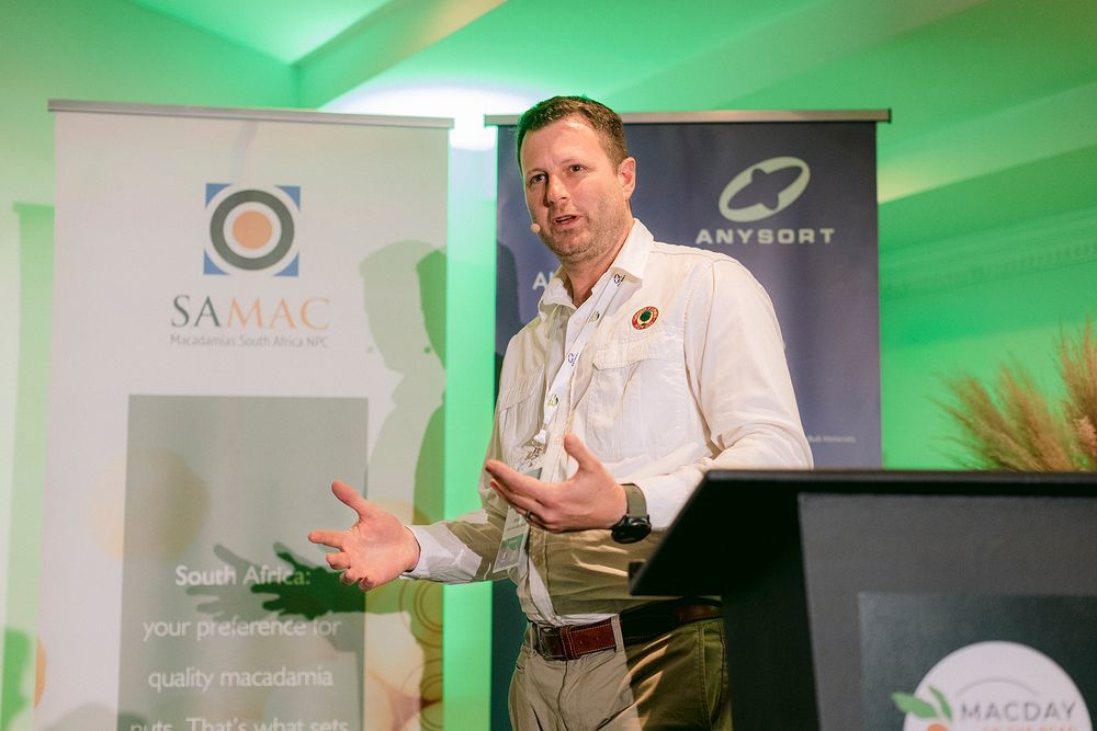 Man presenting at SAMAC and Anysort macadamia industry conference, speaking at podium with branded banners.