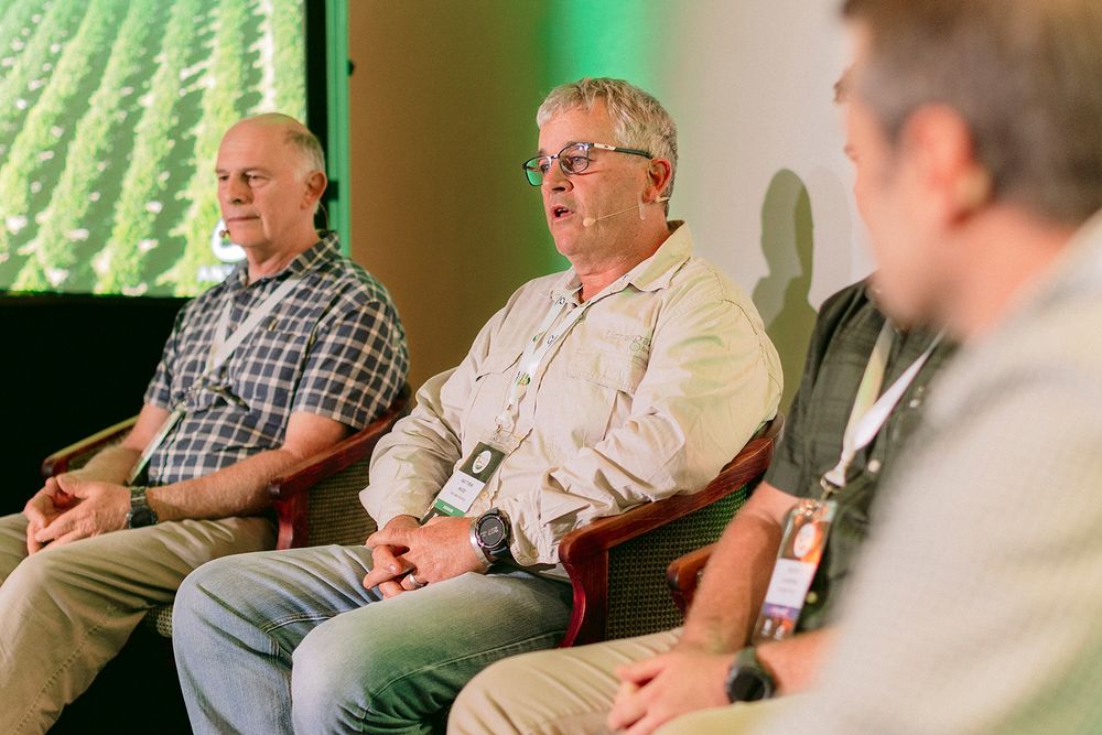 Three men seated on stage during an agricultural panel discussion with green crop field backdrop.