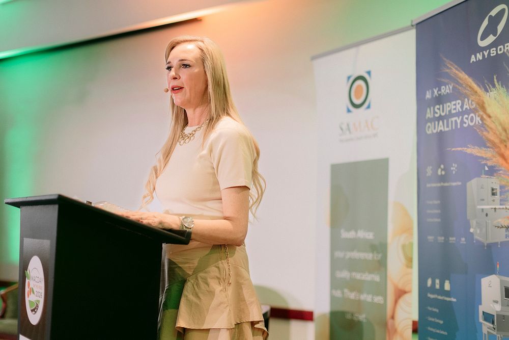 Woman with blonde hair speaking at a podium during a professional conference with branded banners in background.