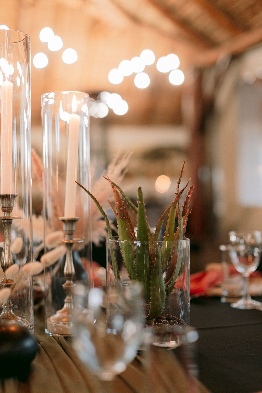Elegant wedding table decor with glass candle holders, aloe vera centerpiece, and bokeh string lights in a rustic barn venue.