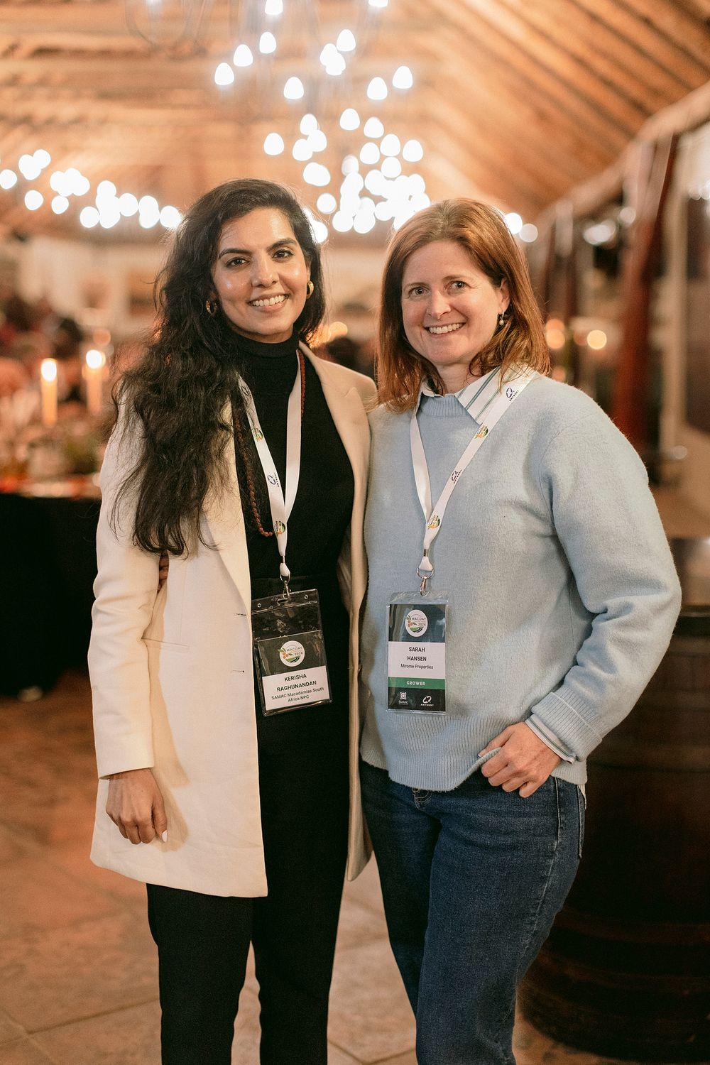 Two women wearing conference lanyards smile together at an indoor event venue with warm string lights.