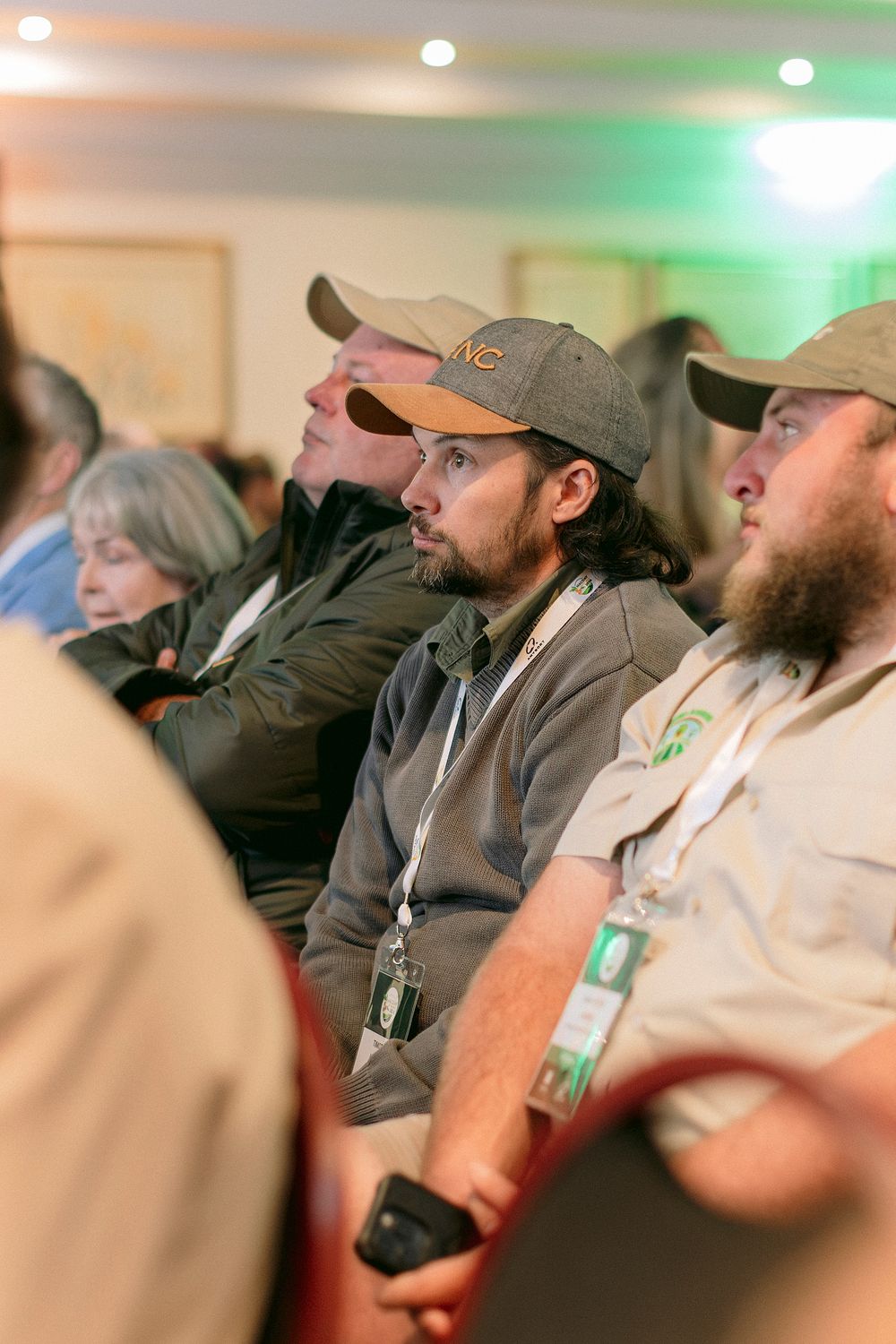 Audience members wearing caps and lanyards attentively watching a presentation at an indoor conference event.
