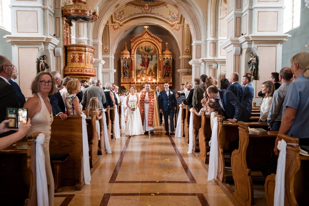 Wedding ceremony in an ornate church with couple walking down the aisle past seated guests.