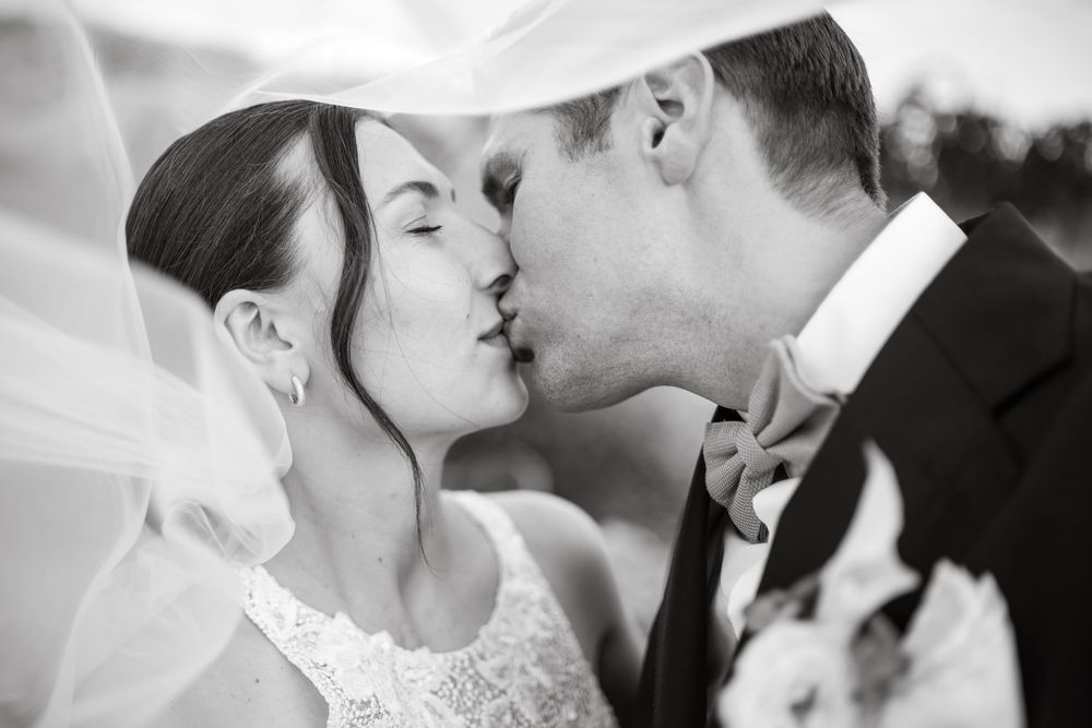 Black and white photo of a bride and groom sharing a romantic kiss under a flowing veil on their wedding day.