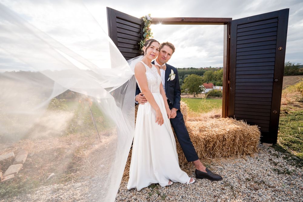 Bride and groom pose outdoors near rustic wooden doors and hay bales, wedding veil flowing in the wind.