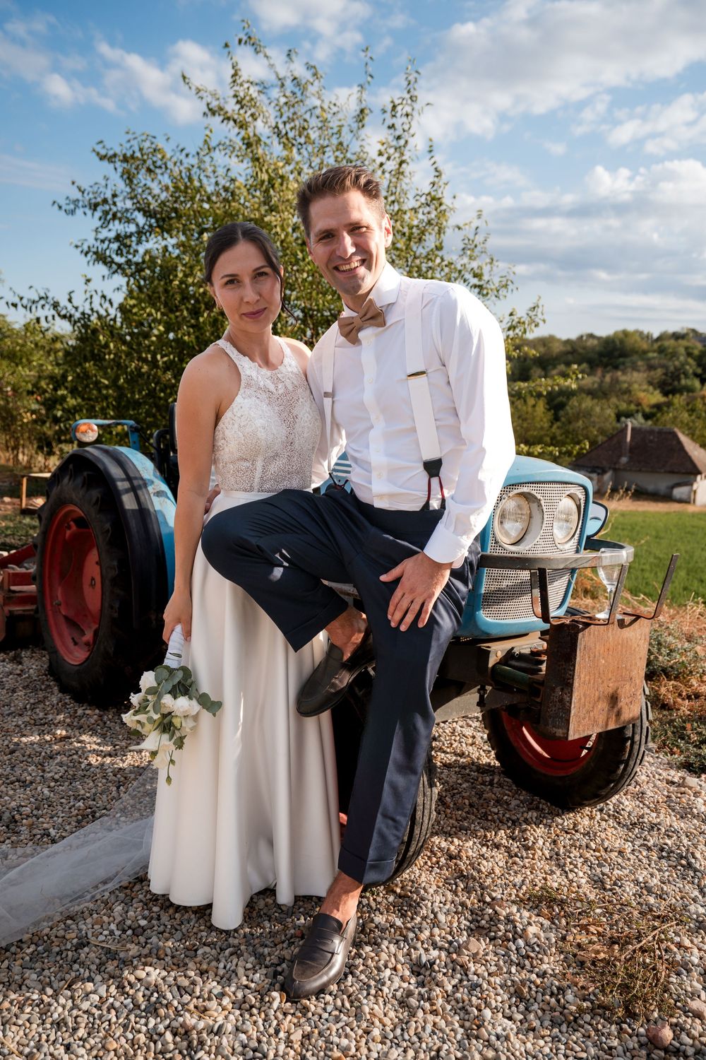 Bride and groom pose together in front of a vintage blue tractor on a sunny wedding day outdoors.