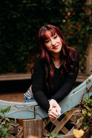 Young woman with red-highlighted dark hair in black top and jeans, smiling outdoors on vintage blue bench.