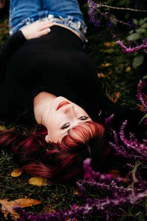Woman with red hair lying on grass surrounded by purple wildflowers in an autumn garden setting.