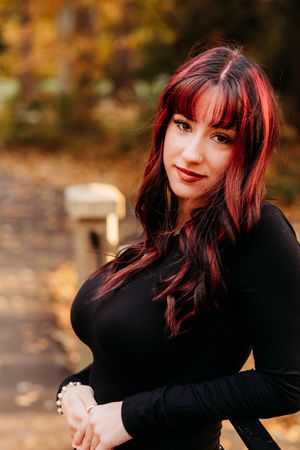 Young woman with red and black hair wearing a black outfit poses outdoors in an autumn park setting.
