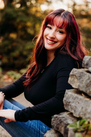 Smiling young woman with red hair wearing a black top and jeans, sitting by a stone wall outdoors.