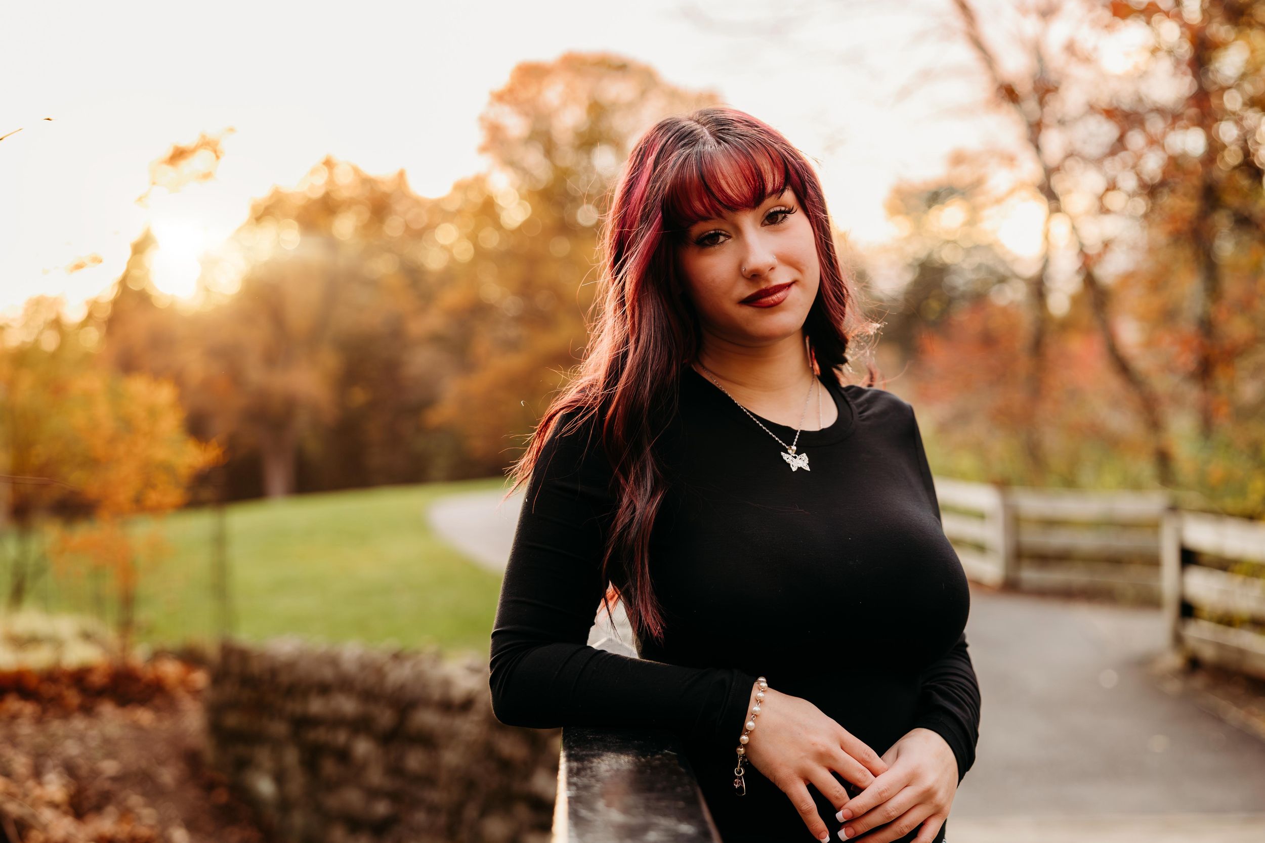 Young woman with red hair in black top poses outdoors during golden hour fall foliage backdrop.
