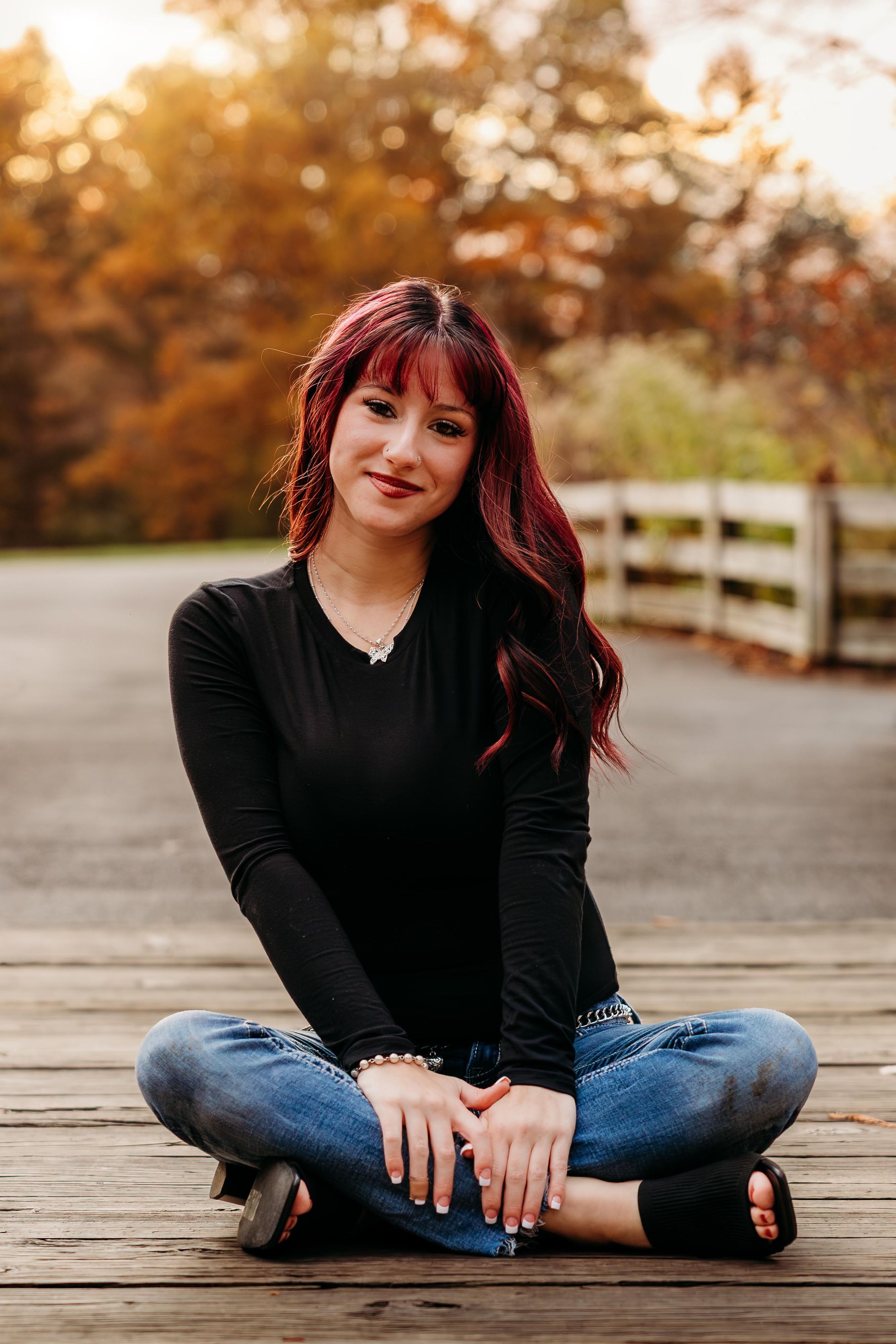 Young woman with red hair sits cross-legged on wooden bridge deck, wearing black top and jeans, autumn trees in background.