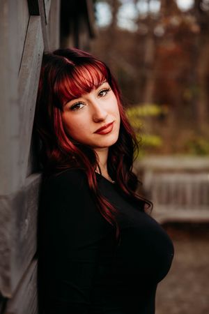 Young woman with red hair and bangs leans against a wooden wall outdoors, wearing a black top.