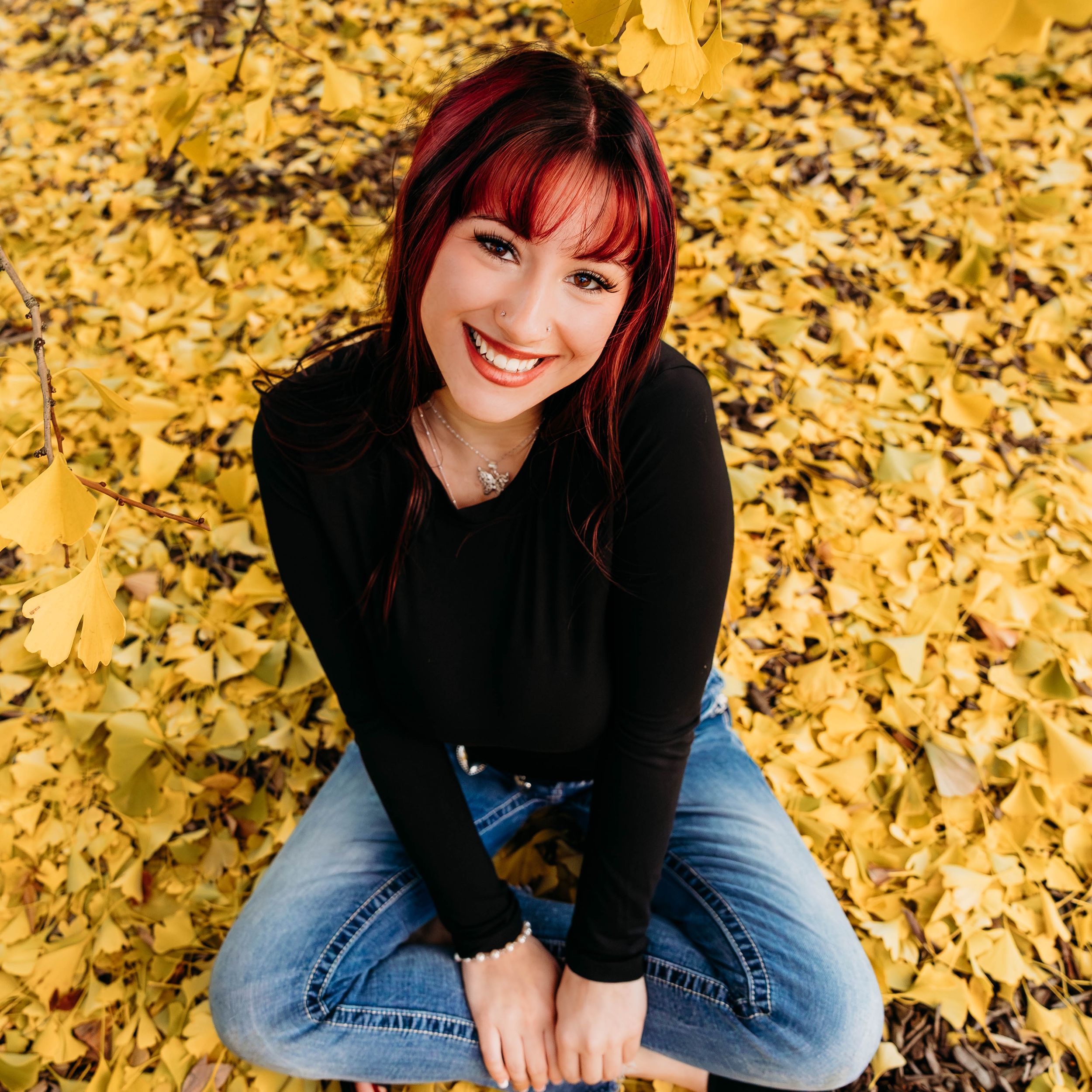 A smiling young woman with red hair sits cross-legged on a bed of golden autumn leaves wearing a black top and jeans.