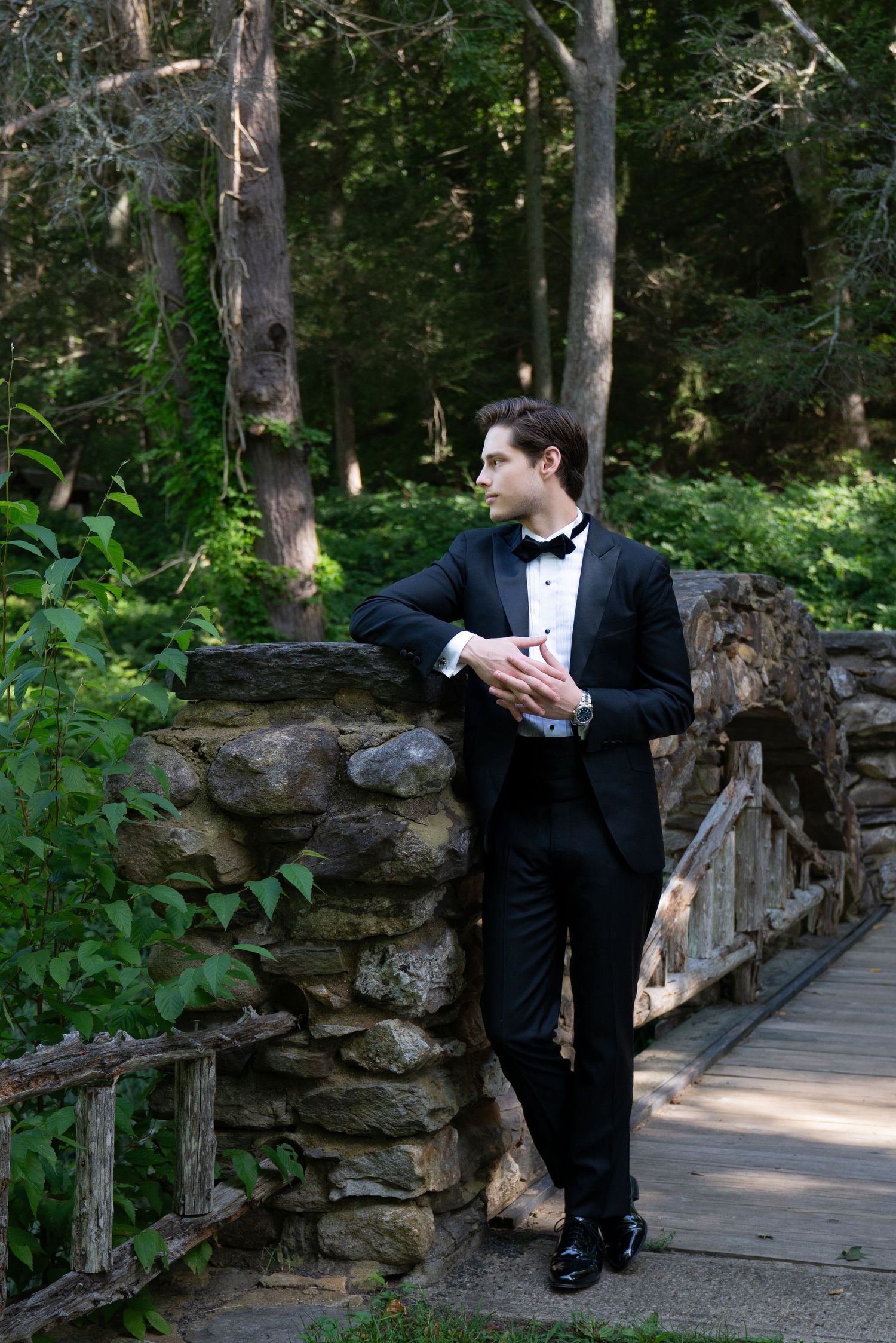 Elegant groom in black tuxedo standing by stone bridge in lush green forest setting.