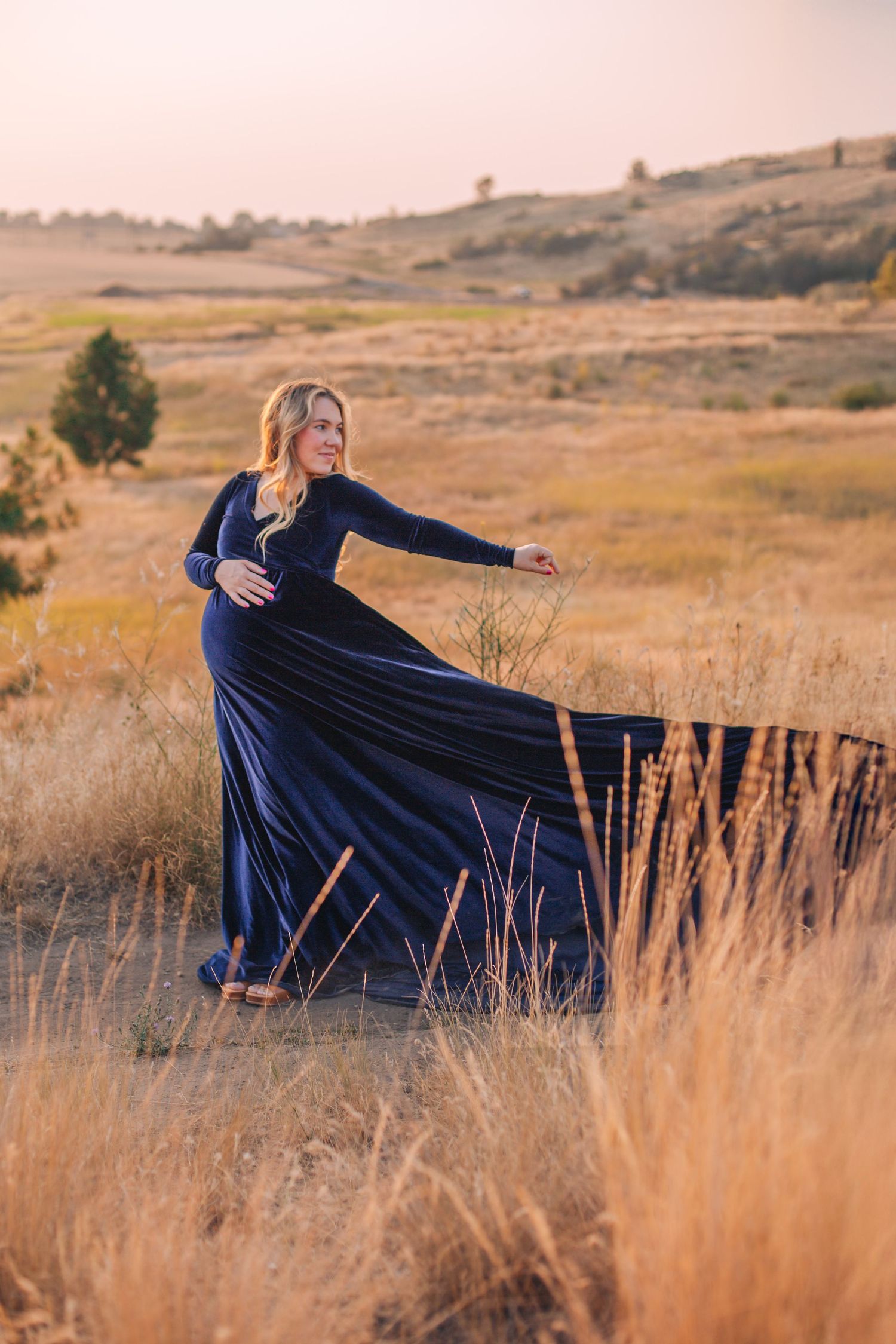 Pregnant woman in flowing navy blue velvet gown posing gracefully in golden field at sunset.