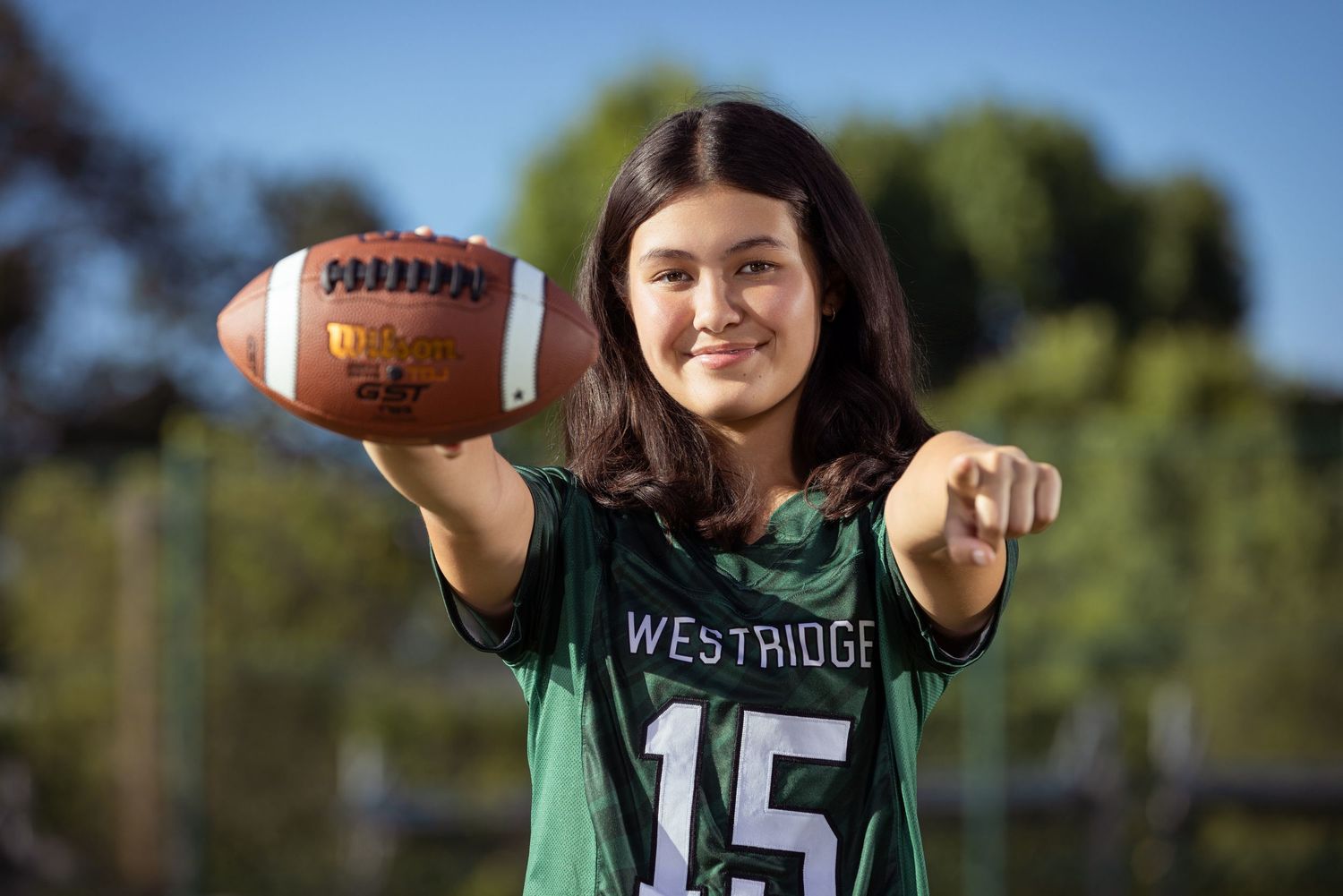 Girl in Westridge #15 jersey holds football and points at camera on outdoor field.