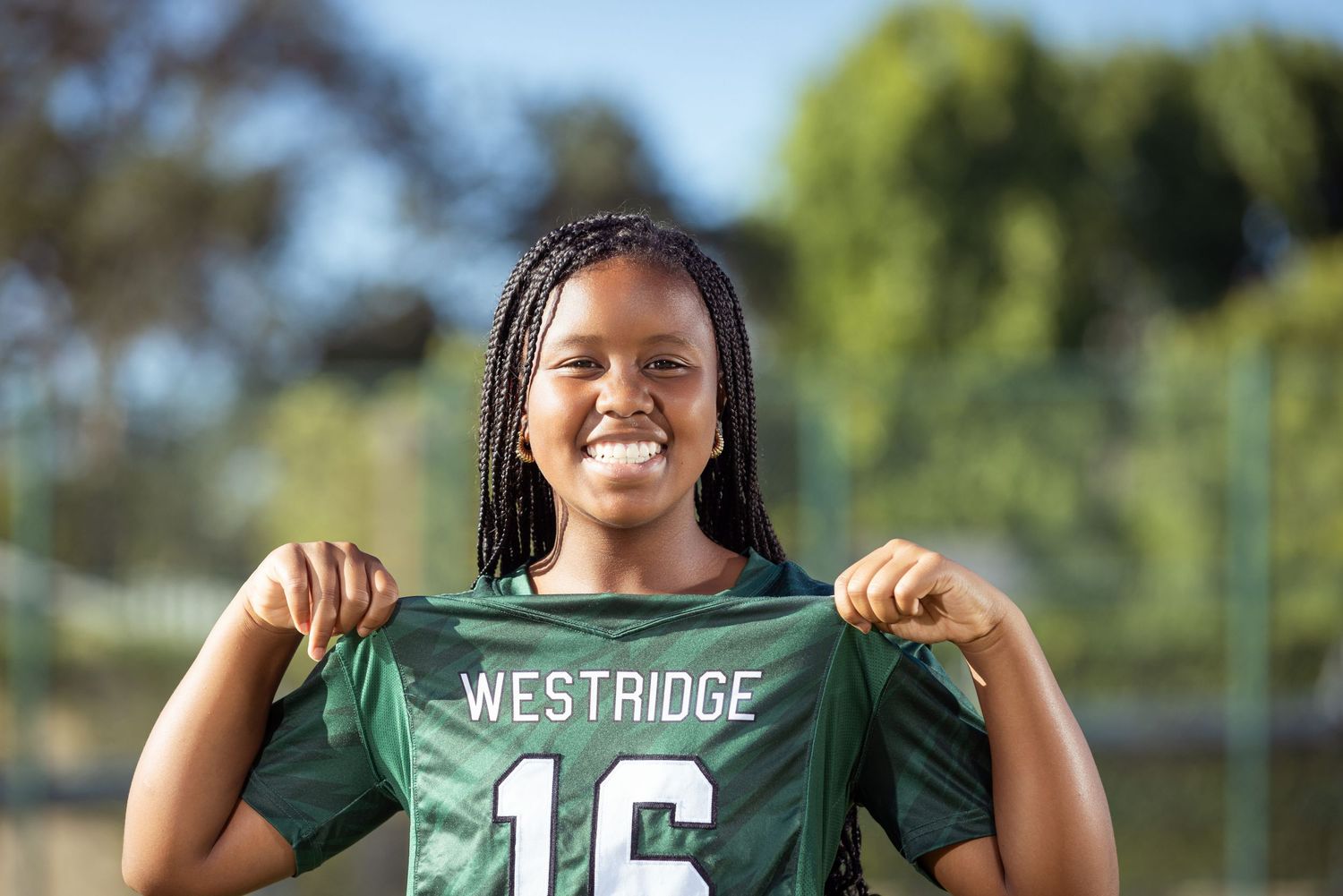 Young female athlete smiling and holding a green Westridge #16 jersey on a sunny outdoor sports field.