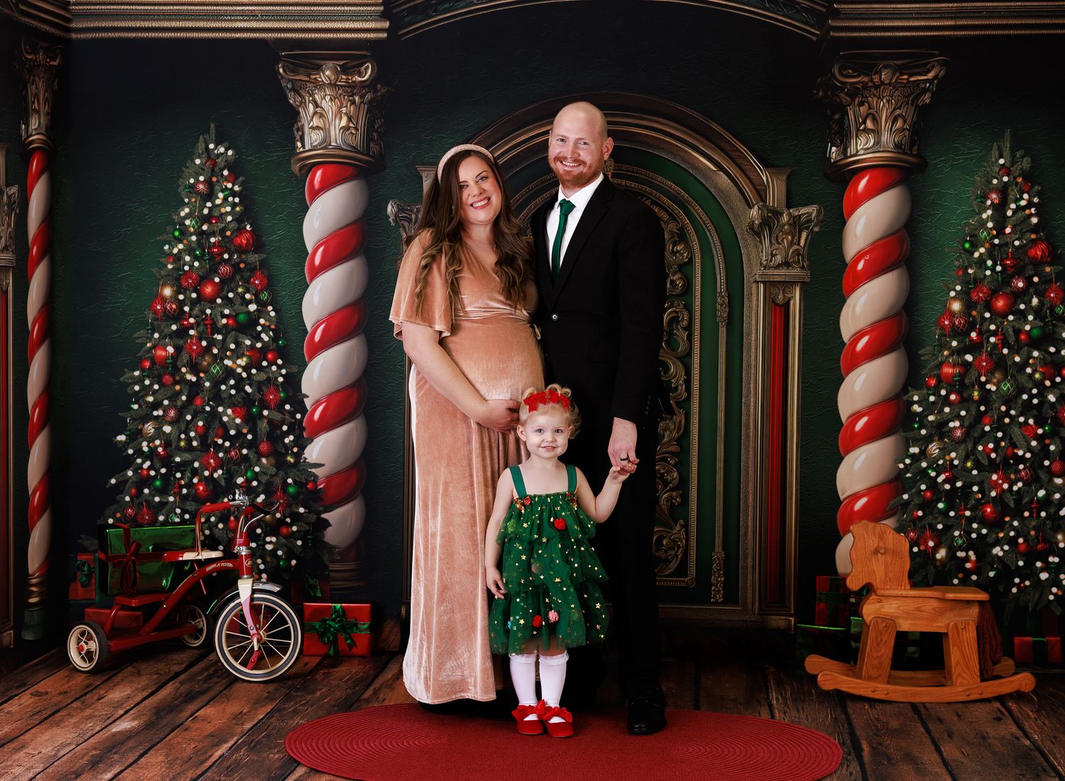 Family Christmas portrait with pregnant mother, father, and toddler girl in festive holiday studio setting with decorated trees.