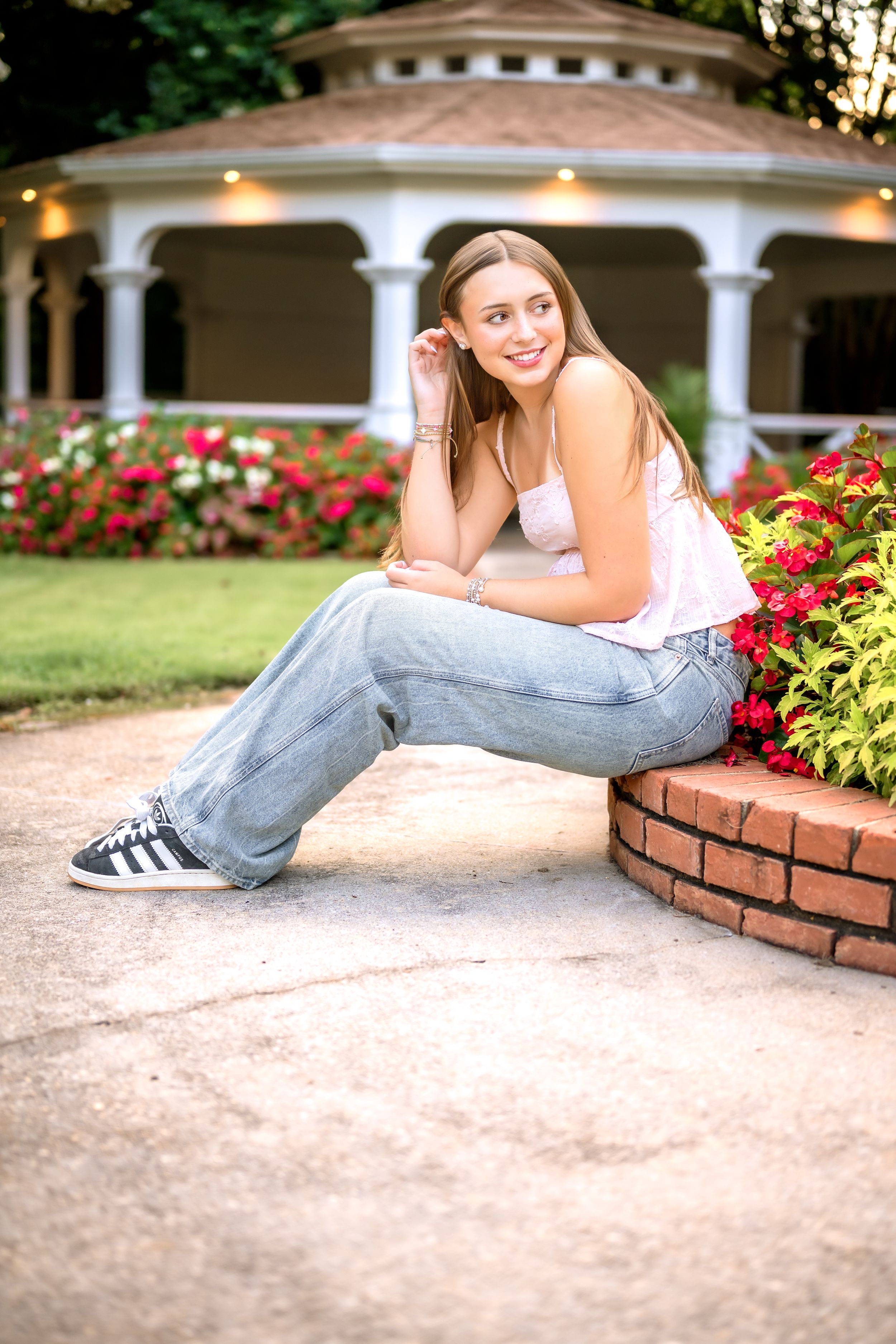 Young woman in jeans and white top sits by brick flower bed, smiling, with white gazebo and red flowers behind her.