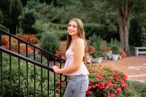 Young woman in white tank top and jeans smiling beside black iron railing with colorful garden flowers in background.