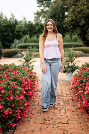 Young woman in pink top and wide-leg jeans walking on brick path lined with red flowers in a garden.