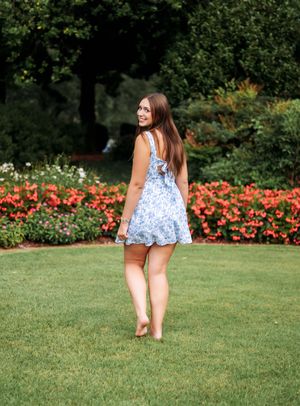 Young woman in blue floral mini dress standing on green lawn with colorful red flowers in background.