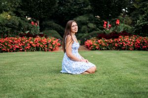 Young woman in blue floral dress sitting on green grass in a garden with red flowers in background.