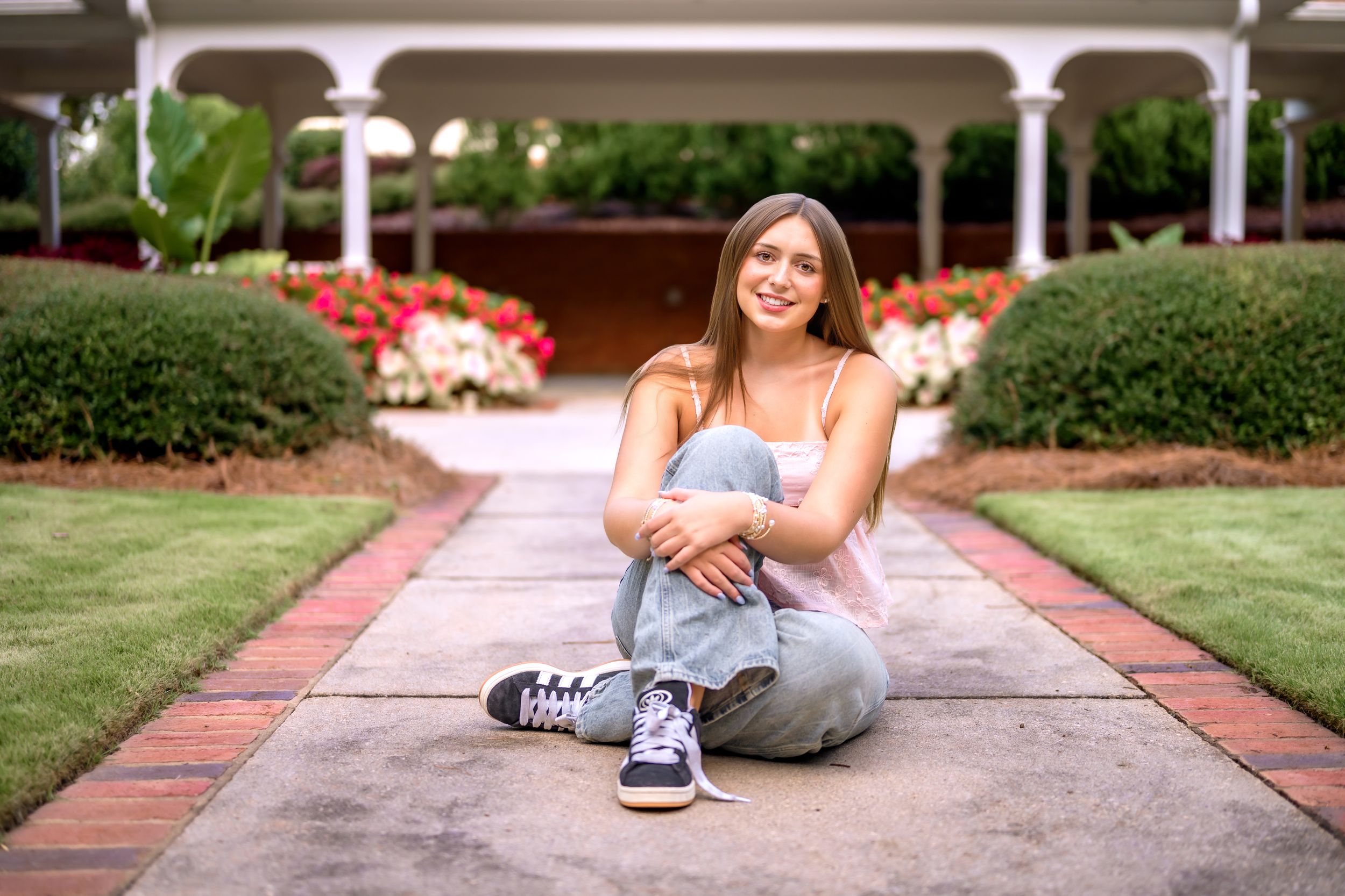 Young woman sitting cross-legged on a garden path, smiling, wearing jeans and sneakers, with colorful flowers in background.