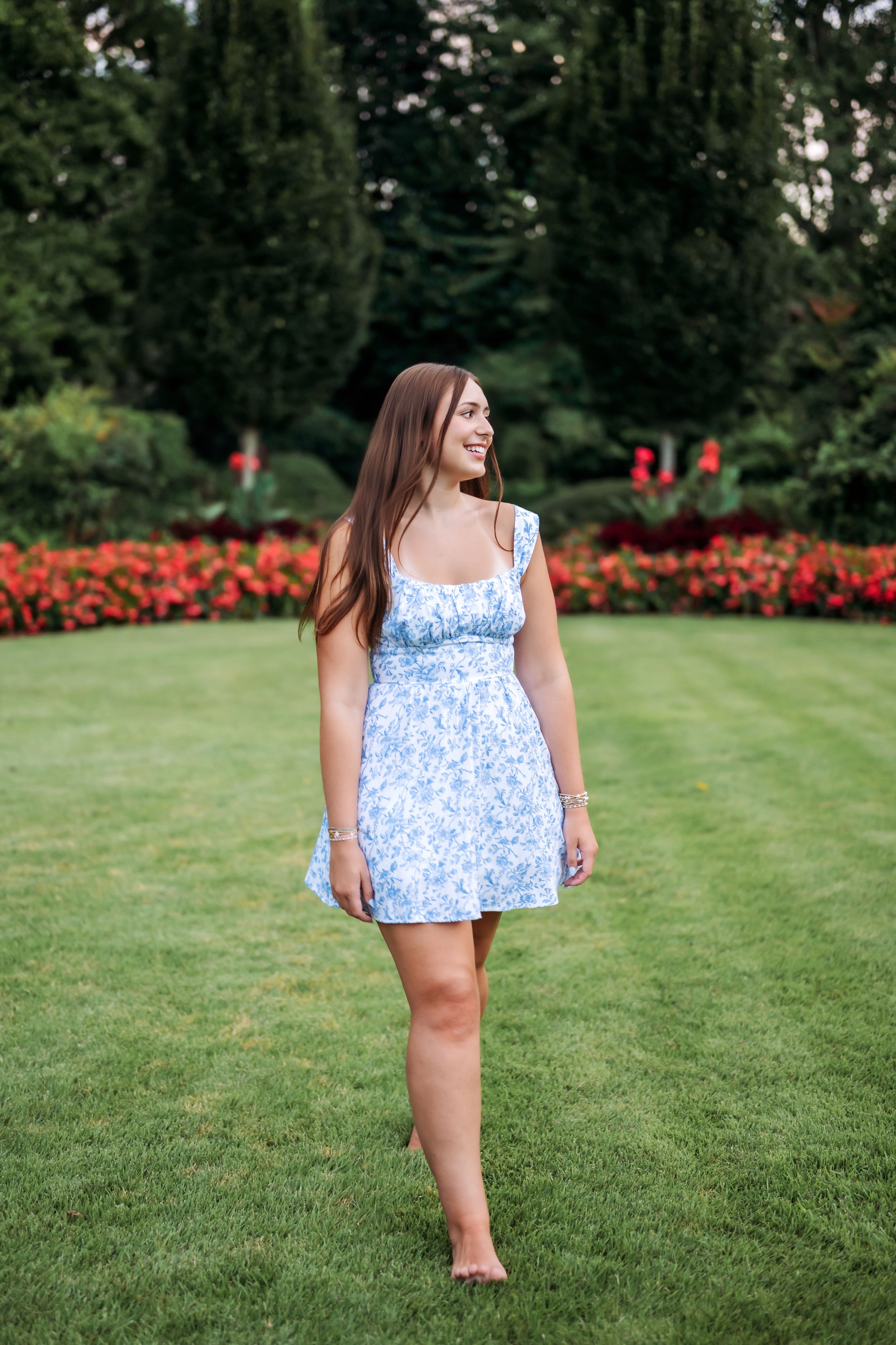 Young woman in blue floral sundress smiling while standing barefoot on green lawn with red flowers and trees.
