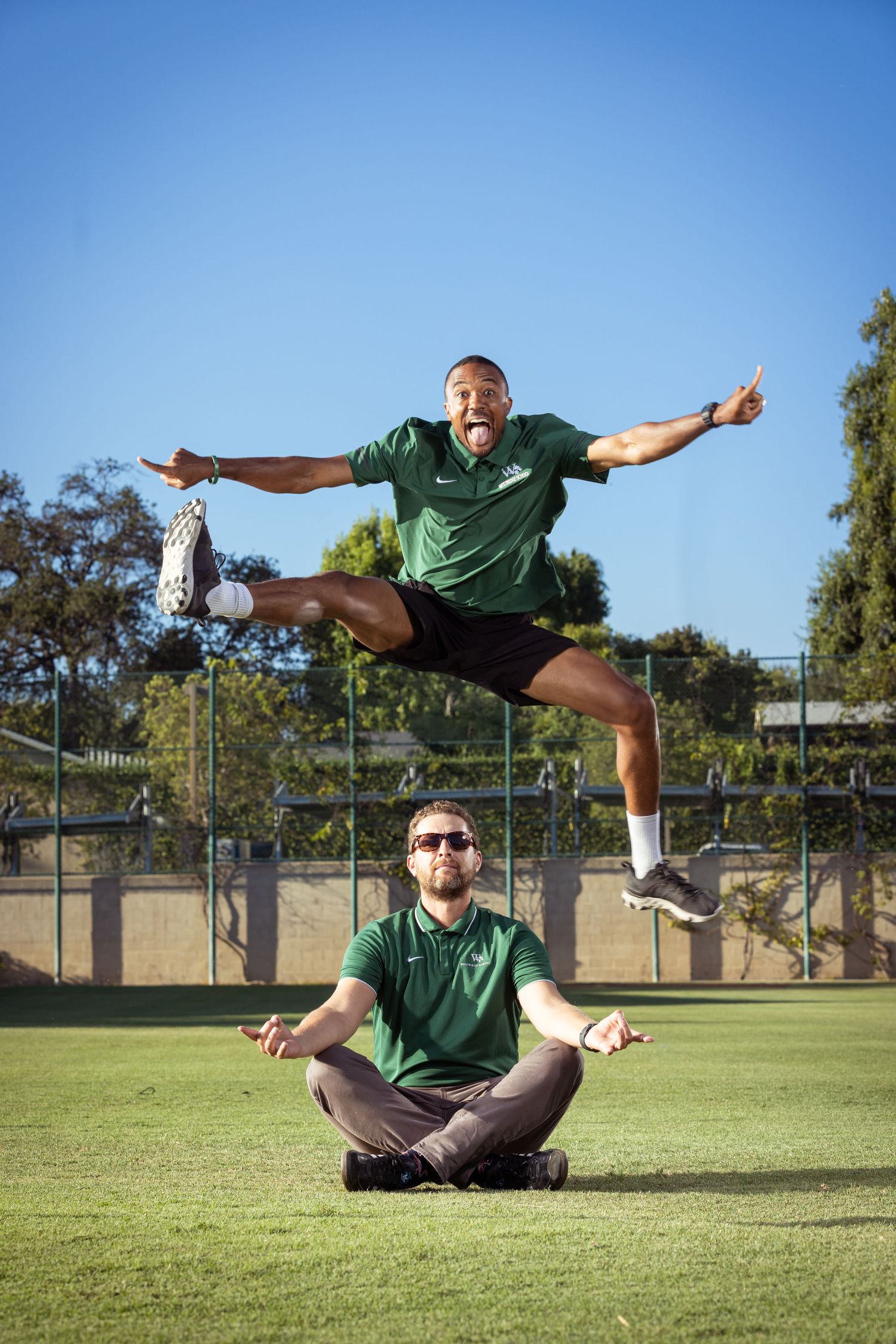 Two men in green shirts on a field, one meditating while the other jumps energetically overhead under a blue sky.