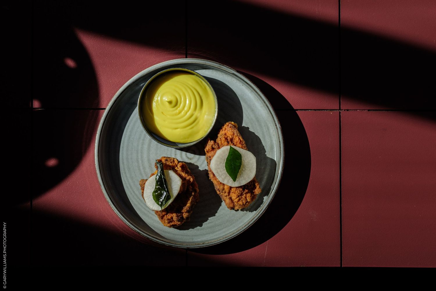 Overhead view of a gourmet plate with whipped yellow sauce, fried bites, and fresh herb garnish on a red tiled surface.
