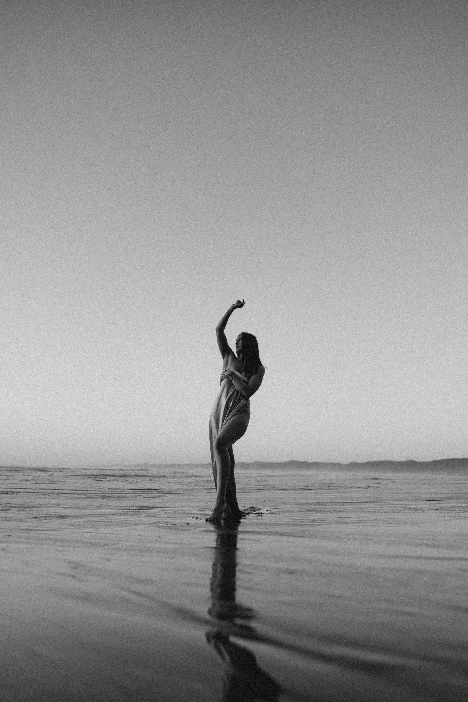 Black and white photo of a woman in a flowing dress standing in shallow water with one arm raised toward the sky.
