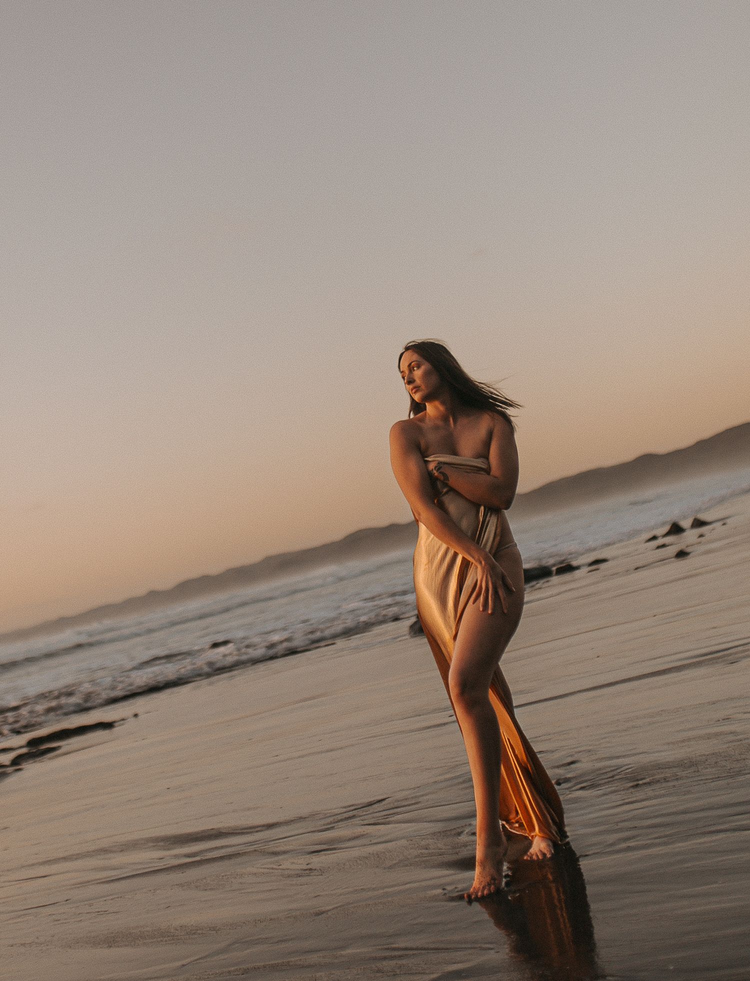 Woman in flowing gold dress standing on black sand beach, Raglan New Zealand