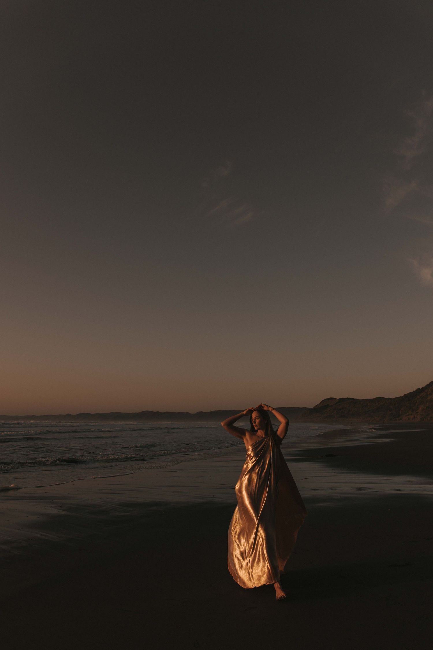 Woman draped in gold fabric on Raglan's black sand beach at sunset — Goddess Shoot® by Nami Creative