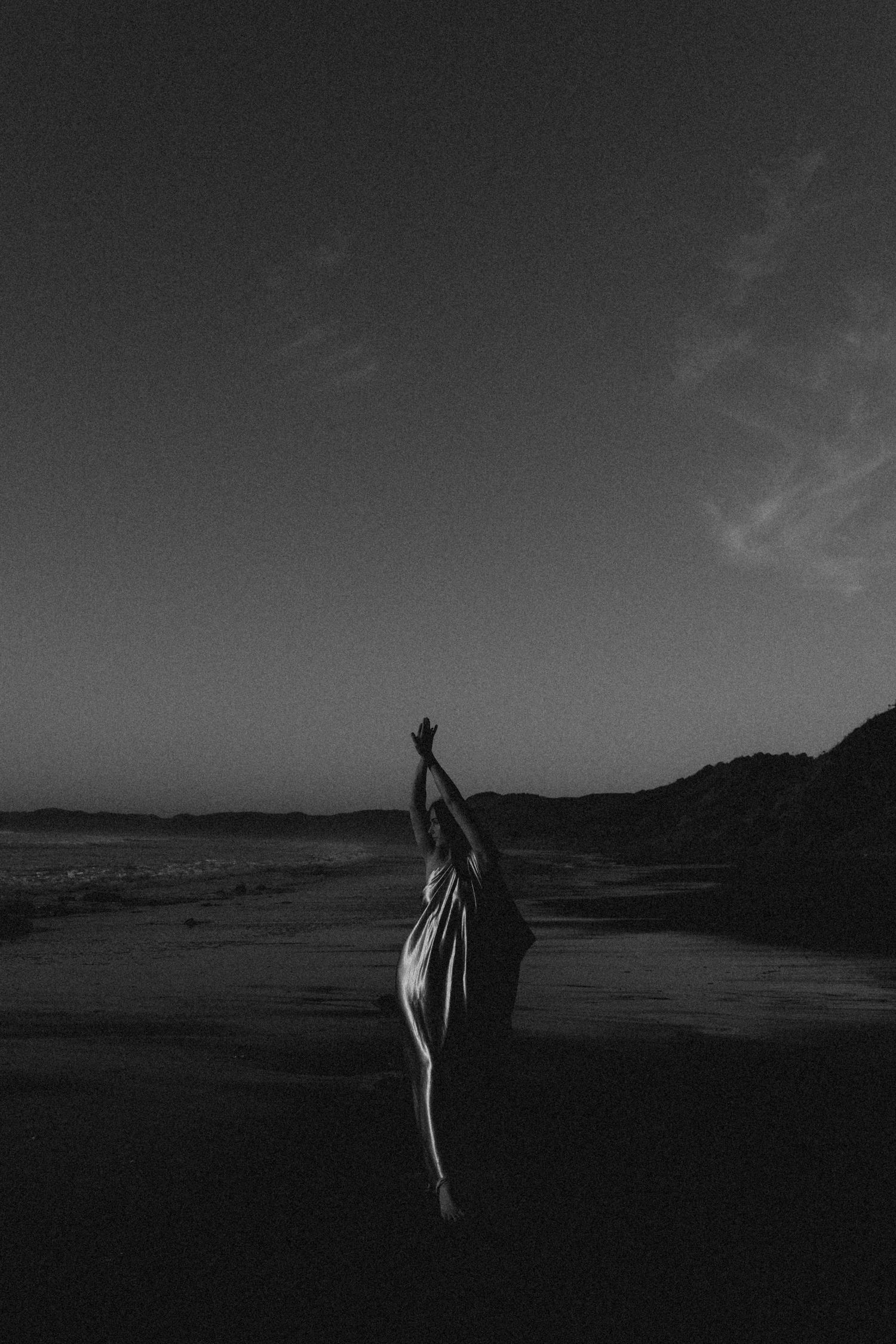 Black and white photo of a woman in a dress reaching arms up toward the sky on a dark beach at dusk.