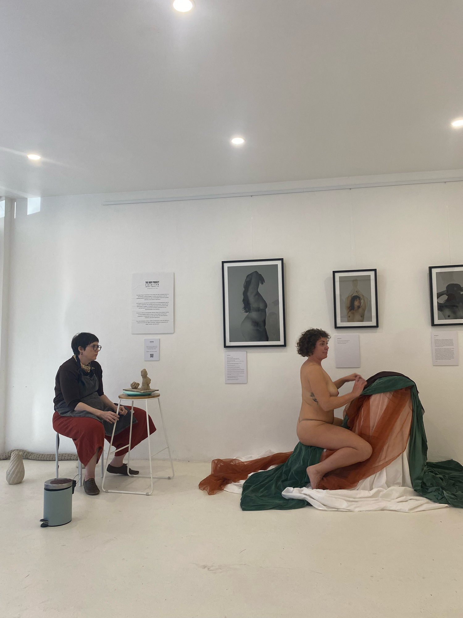 Artist sketching a nude model draped in green and orange fabric in a white gallery with framed artwork on the wall.
