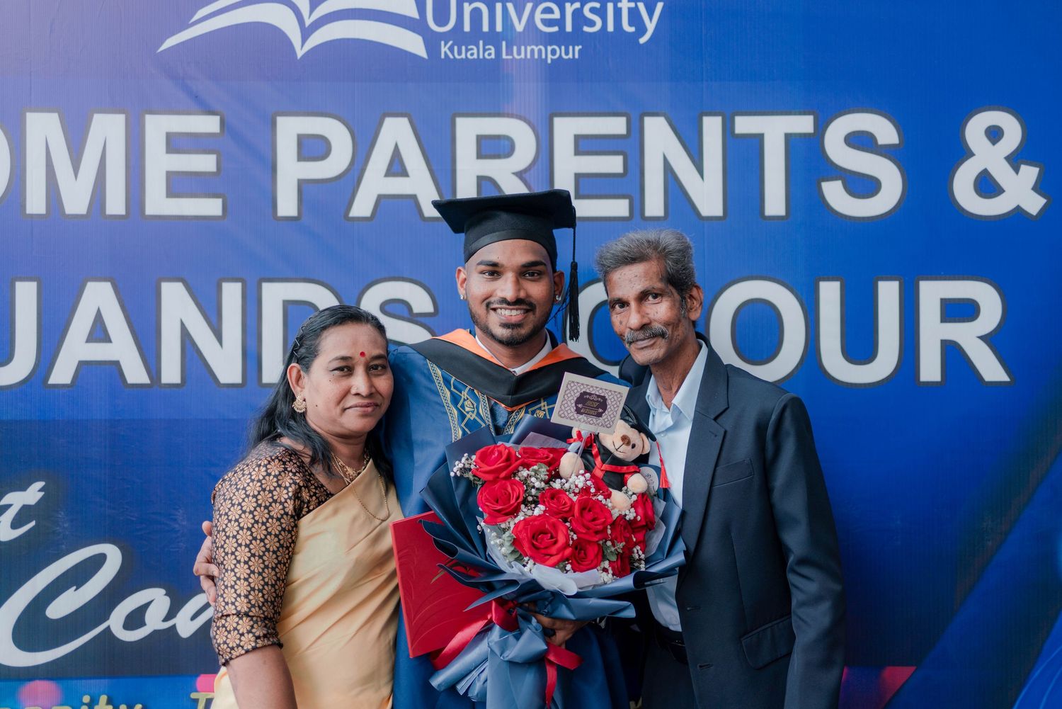 Graduate in cap and gown holds diploma, flanked by two friends with bouquets, posing near a fountain.
