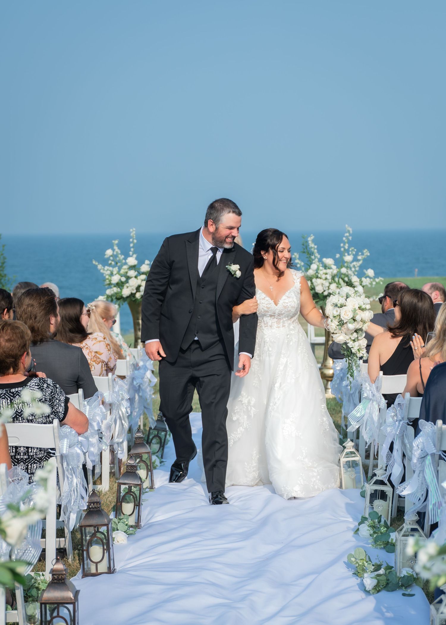 Bride and groom walk down the aisle at an outdoor oceanfront wedding ceremony with white floral arrangements.