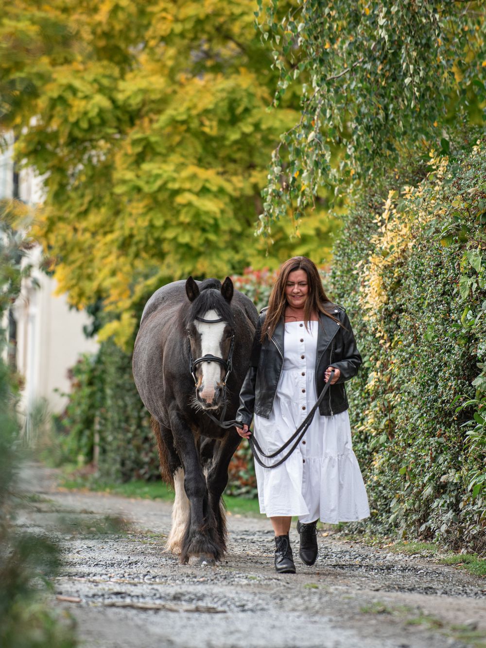 New horse owner Justine wearing a white boho dress with a leather jacket and boots walking down a lane alongside her black miniature cob
