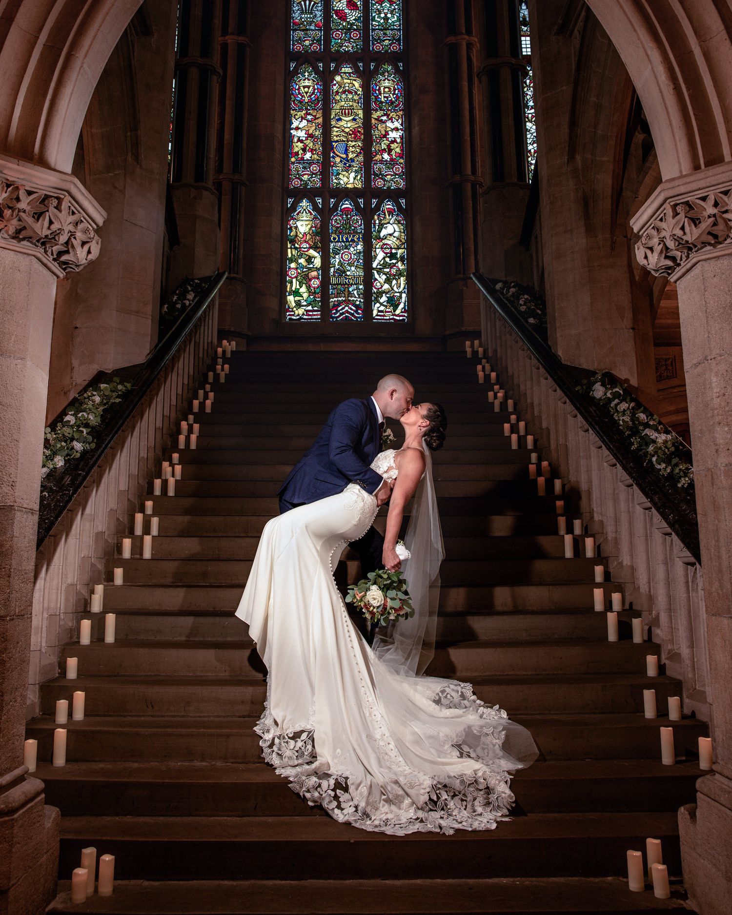 Bride and groom share a kiss on a grand candlelit stone staircase beneath a stained glass window, her lace wedding gown trailing behind.