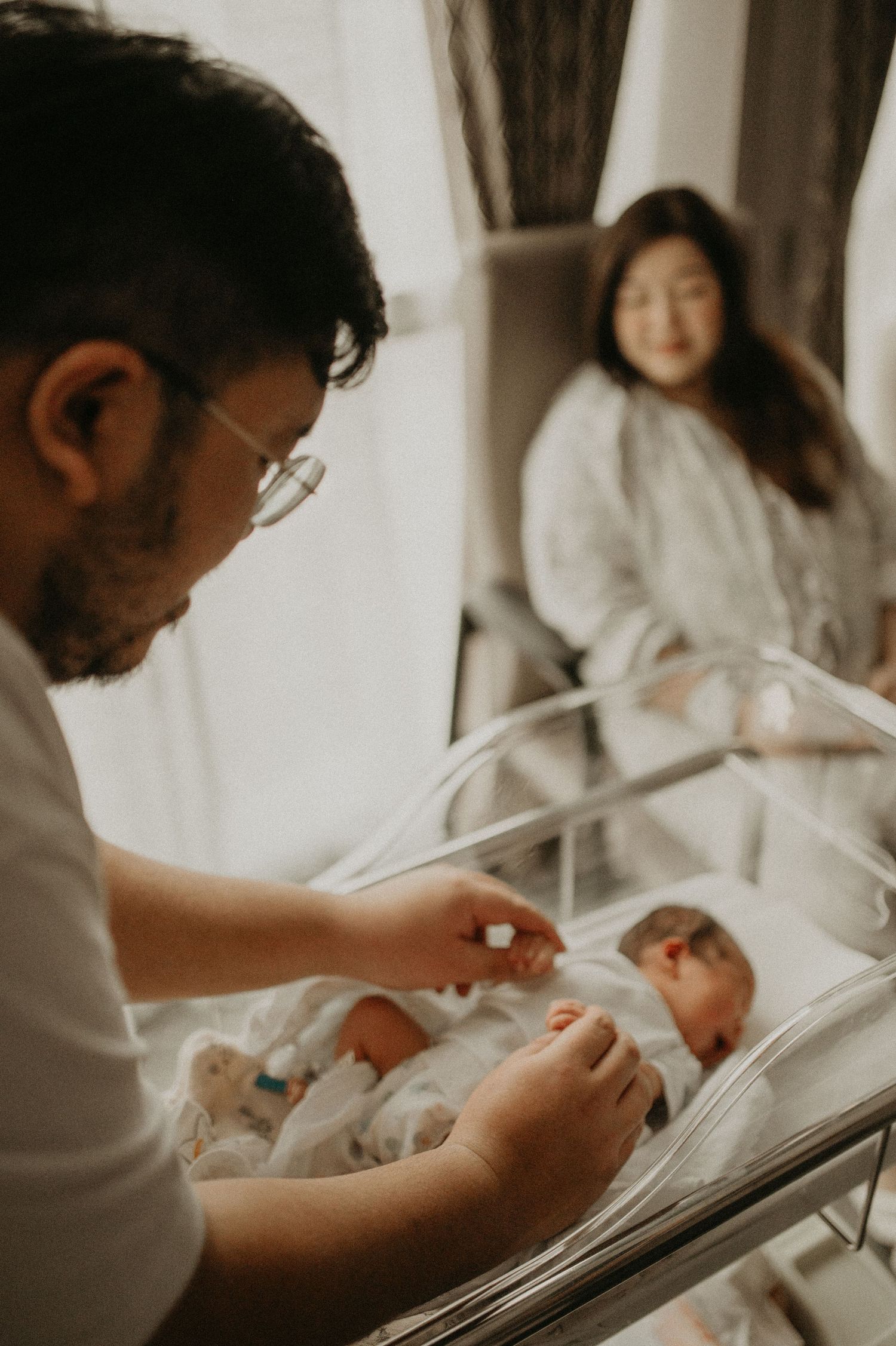 A father in glasses gently touches his newborn baby in a hospital bassinet while the mother rests in a chair in the background.