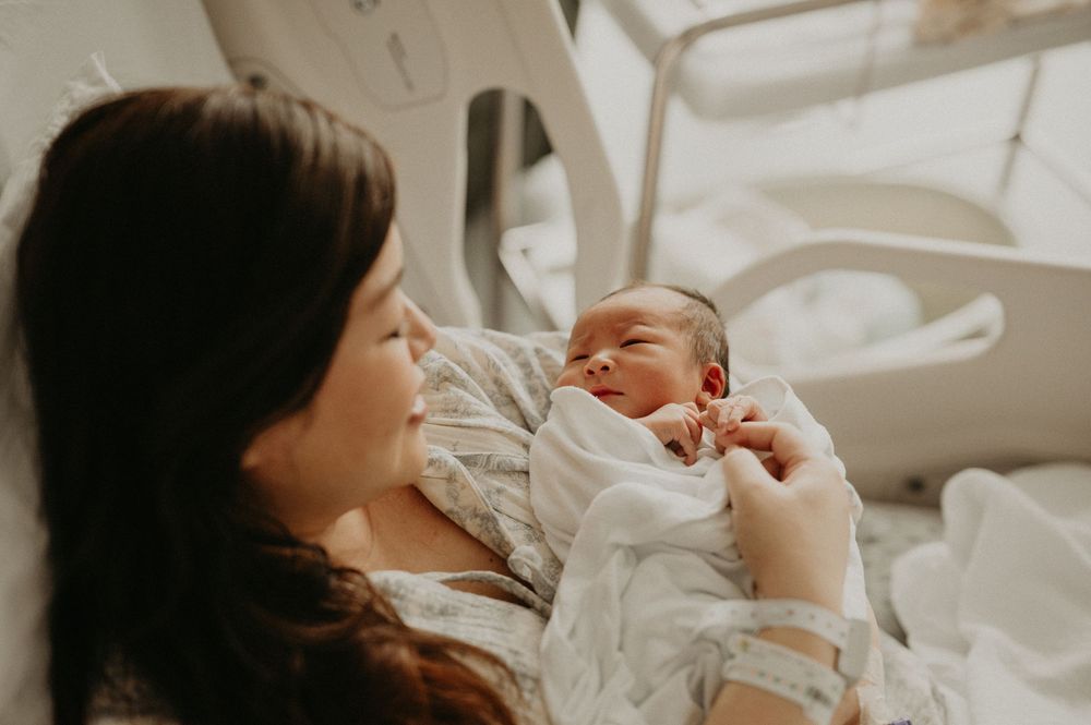 New mother in a hospital bed gently holds her sleeping newborn baby wrapped in a white blanket in a bright delivery room.