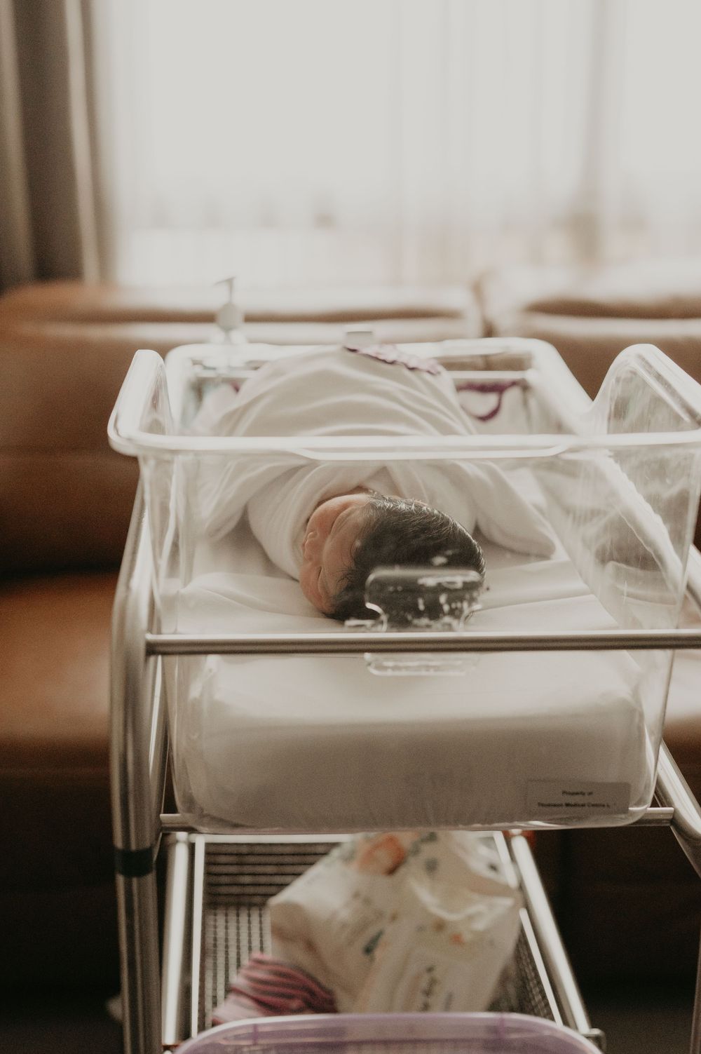 Newborn baby sleeping swaddled in a clear hospital bassinet beside a window in a bright, softly lit maternity room.