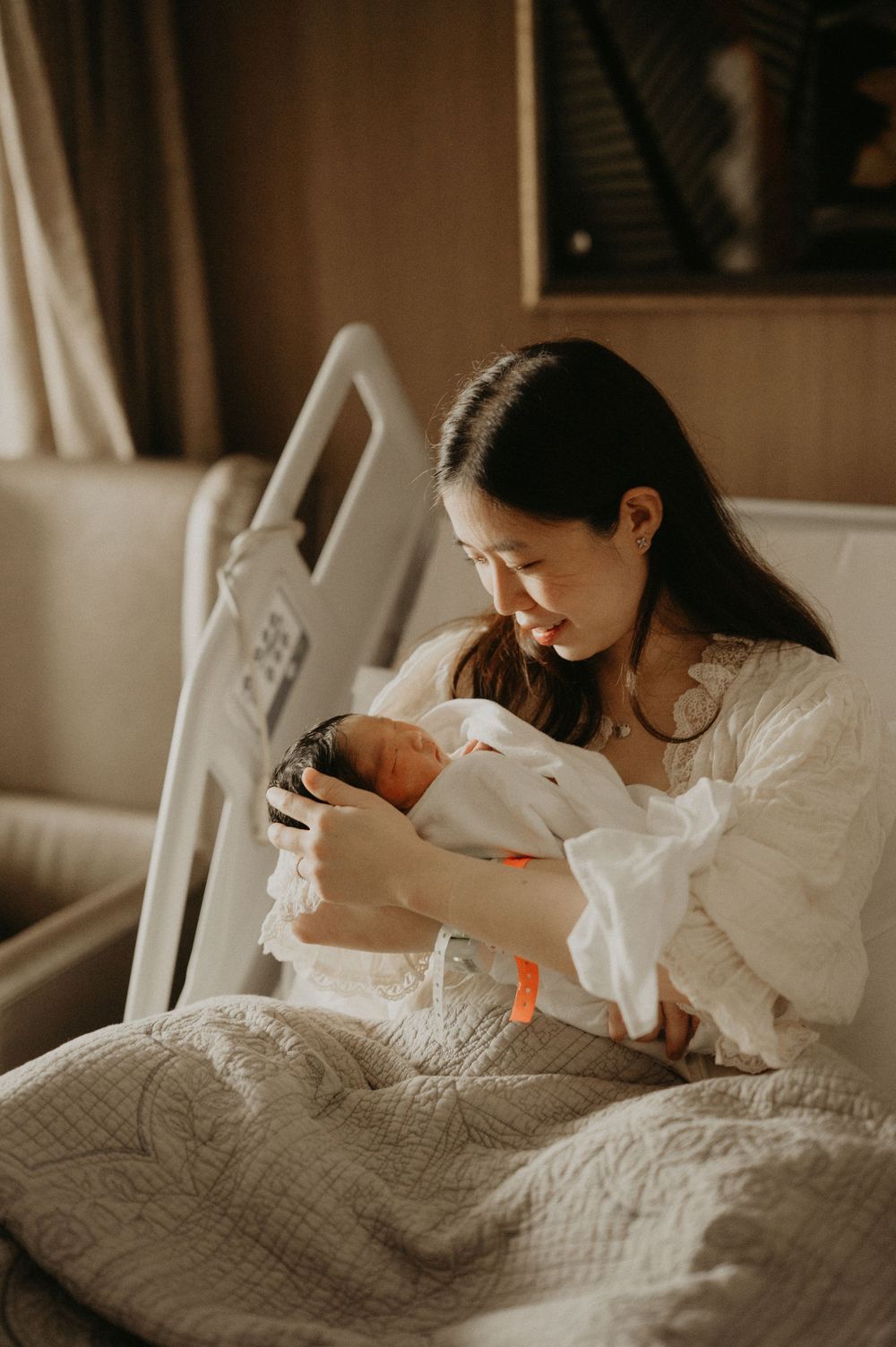 A smiling new mother in a white blouse sits on a hospital bed, gently cradling her sleeping newborn baby wrapped in a soft blanket.