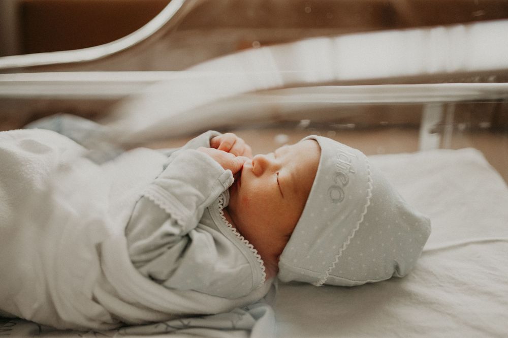 Sleeping newborn baby in a light gray outfit and matching knit cap resting a hand near the mouth in a hospital bassinet.