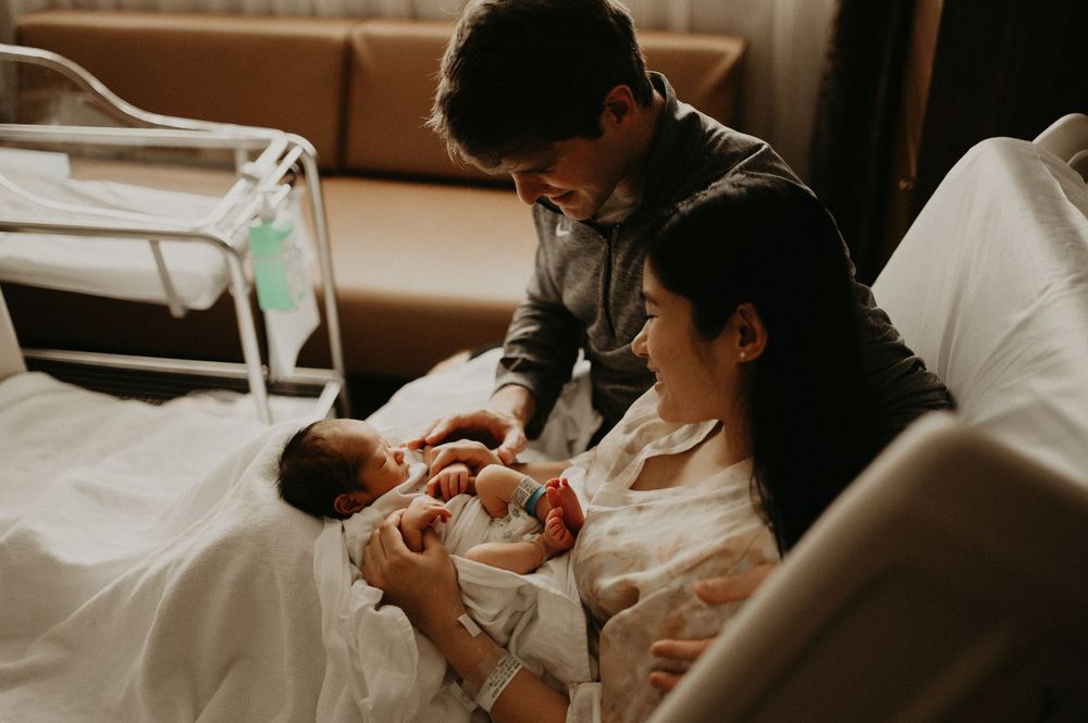 New parents smile down at their newborn baby swaddled in a hospital bed in a warm, softly lit maternity room.