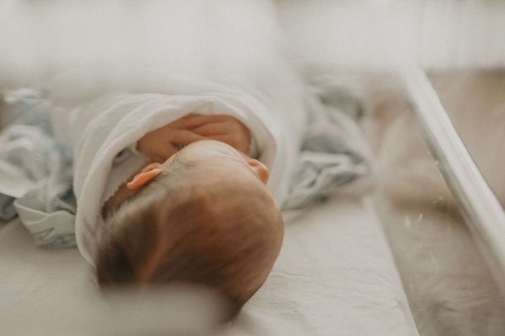 Newborn baby swaddled in a white blanket sleeping peacefully in a hospital bassinet with soft natural light.