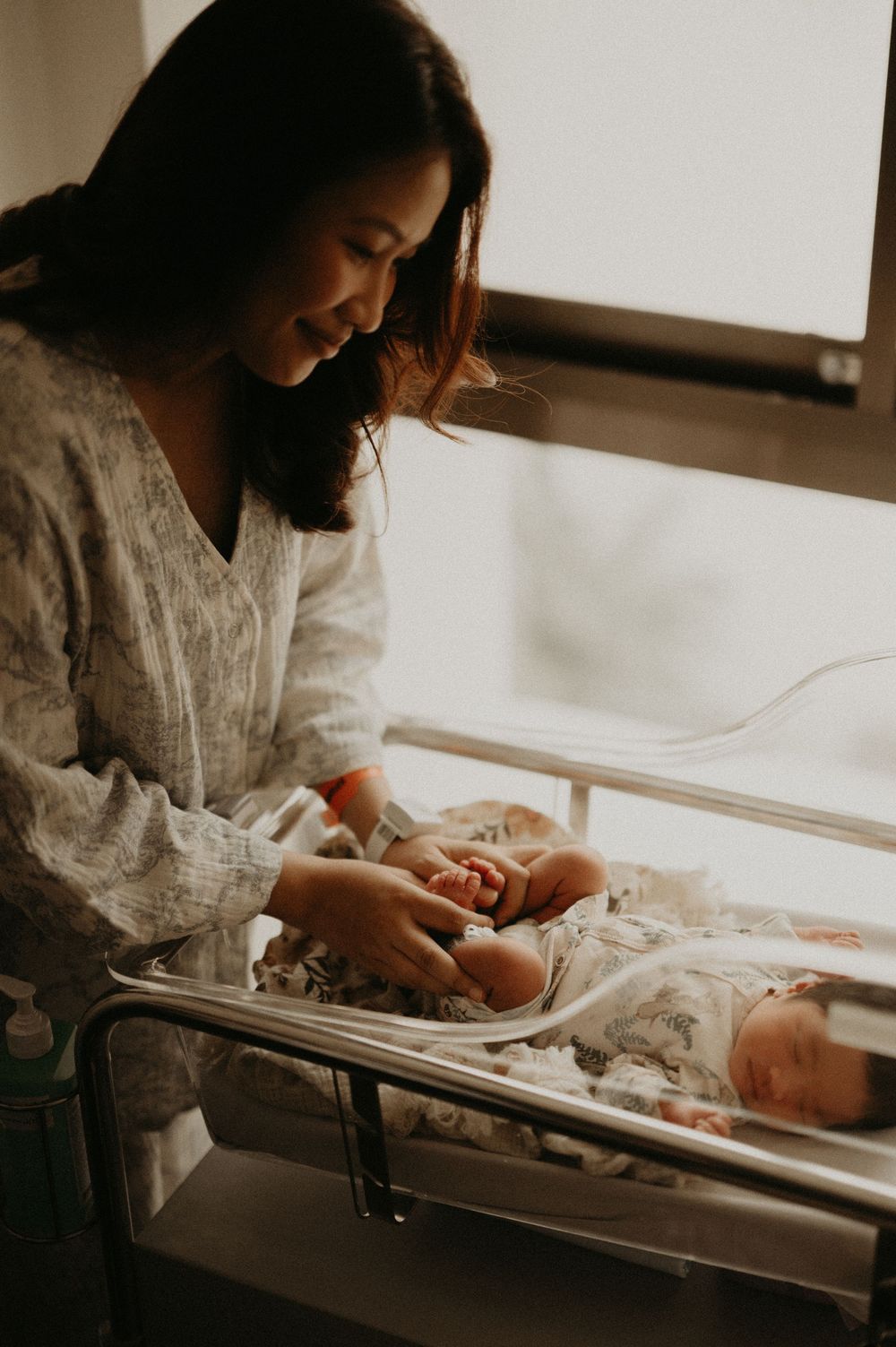 A smiling mother in a patterned hospital gown gently touches her newborn baby sleeping in a clear bassinet beside a sunlit window.